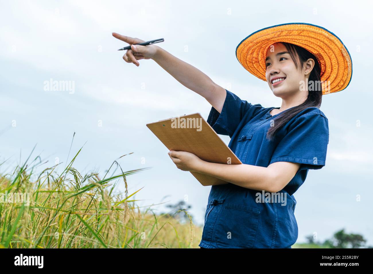 Porträt Asiatische junge Bauernfrau mit Strohhut und Klemmbrett in der Hand, lächelt und zeigt den Finger nach vorne während der Arbeit auf dem Reisfeld Stockfoto
