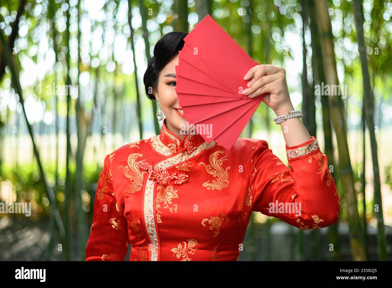 Porträt hübsche asiatische Frau in einem chinesischen Cheongsam, die verstecktes Auge mit roten Umschlägen Fächer im Bambuswald Hintergrund, Kopierraum, chinesisches Neujahr posiert Stockfoto