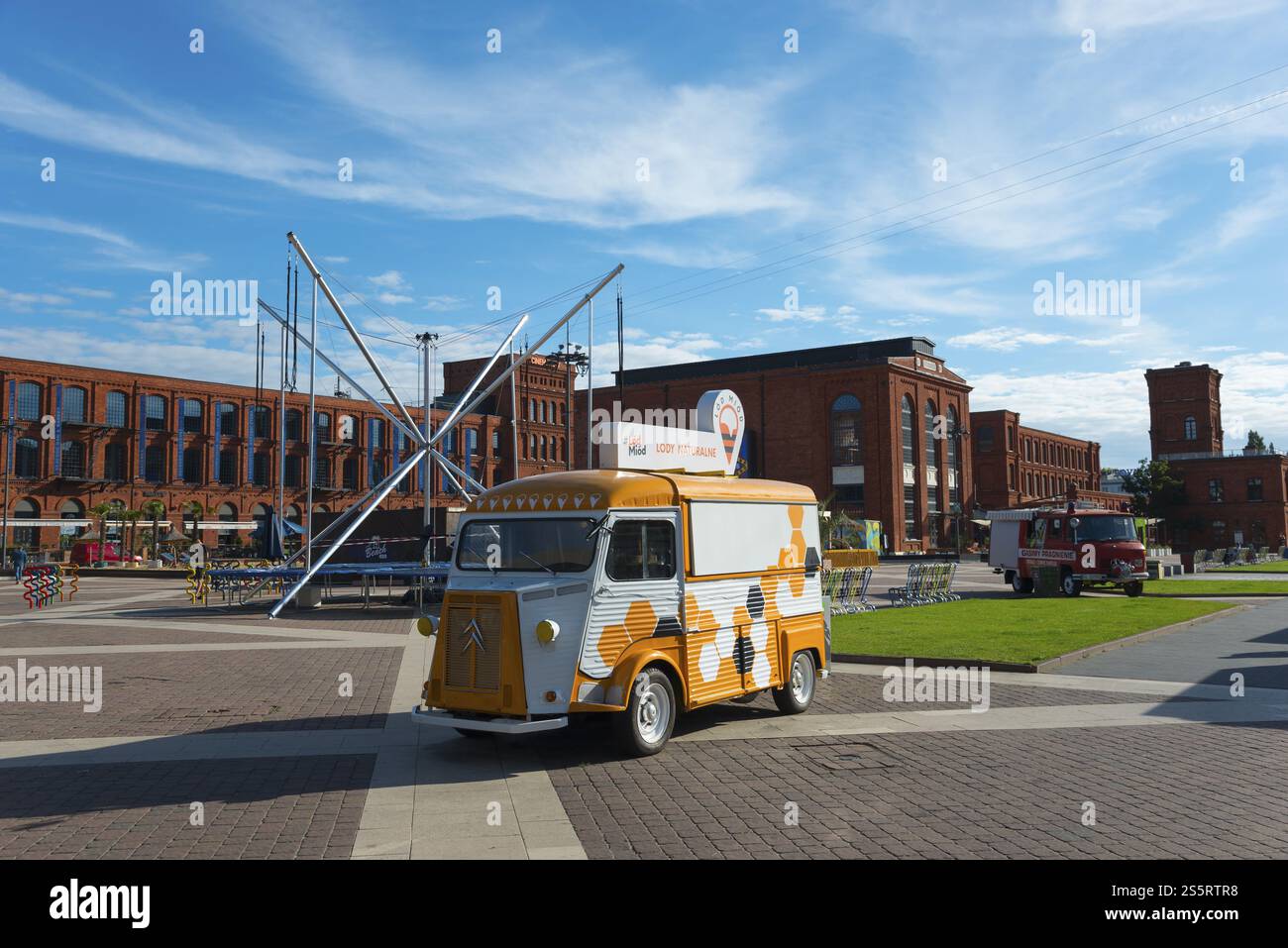 Ein gelb-weißer Retro-Van steht auf einem Stadtplatz vor Industrieziegelbauten und einem Kunstwerk, Manufaktura, Lodz, Lodz, Lodz, Lodz, Polan Stockfoto