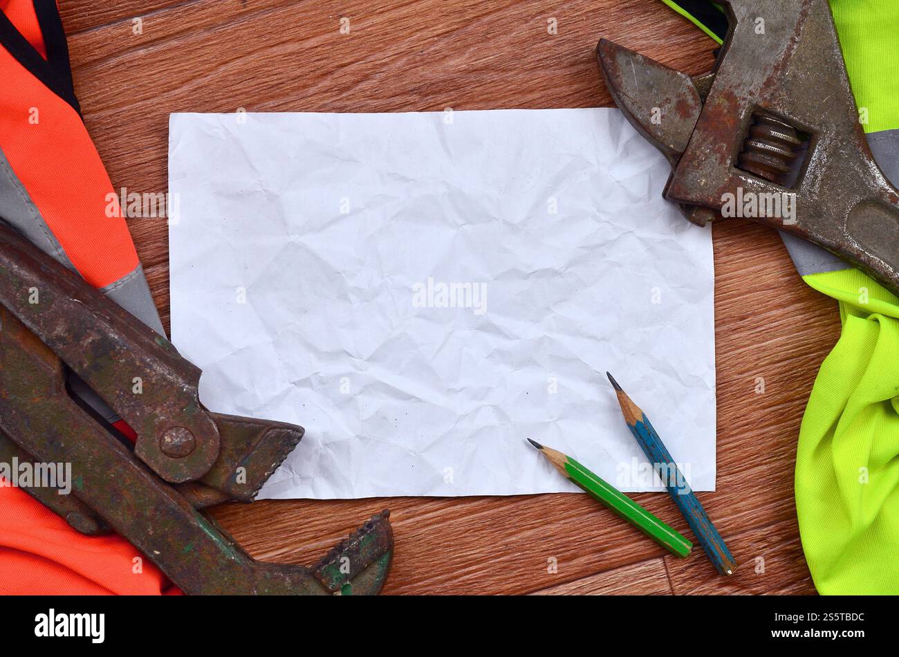 Ein zerknittertes Blatt Papier mit zwei Bleistiften, umgeben von grünen und orangen Arbeitsuniformen und verstellbaren Schraubenschlüsseln. Stillleben in Verbindung mit Stockfoto
