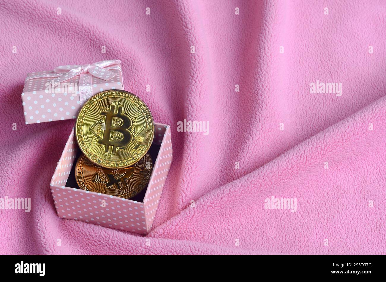Der goldene Bitcoin liegt in einer kleinen rosafarbenen Geschenkbox mit einer kleinen Schleife auf einer Decke aus weichem und flauschigem hellrosa Fleecestoff mit einer großen Anzahl Stockfoto