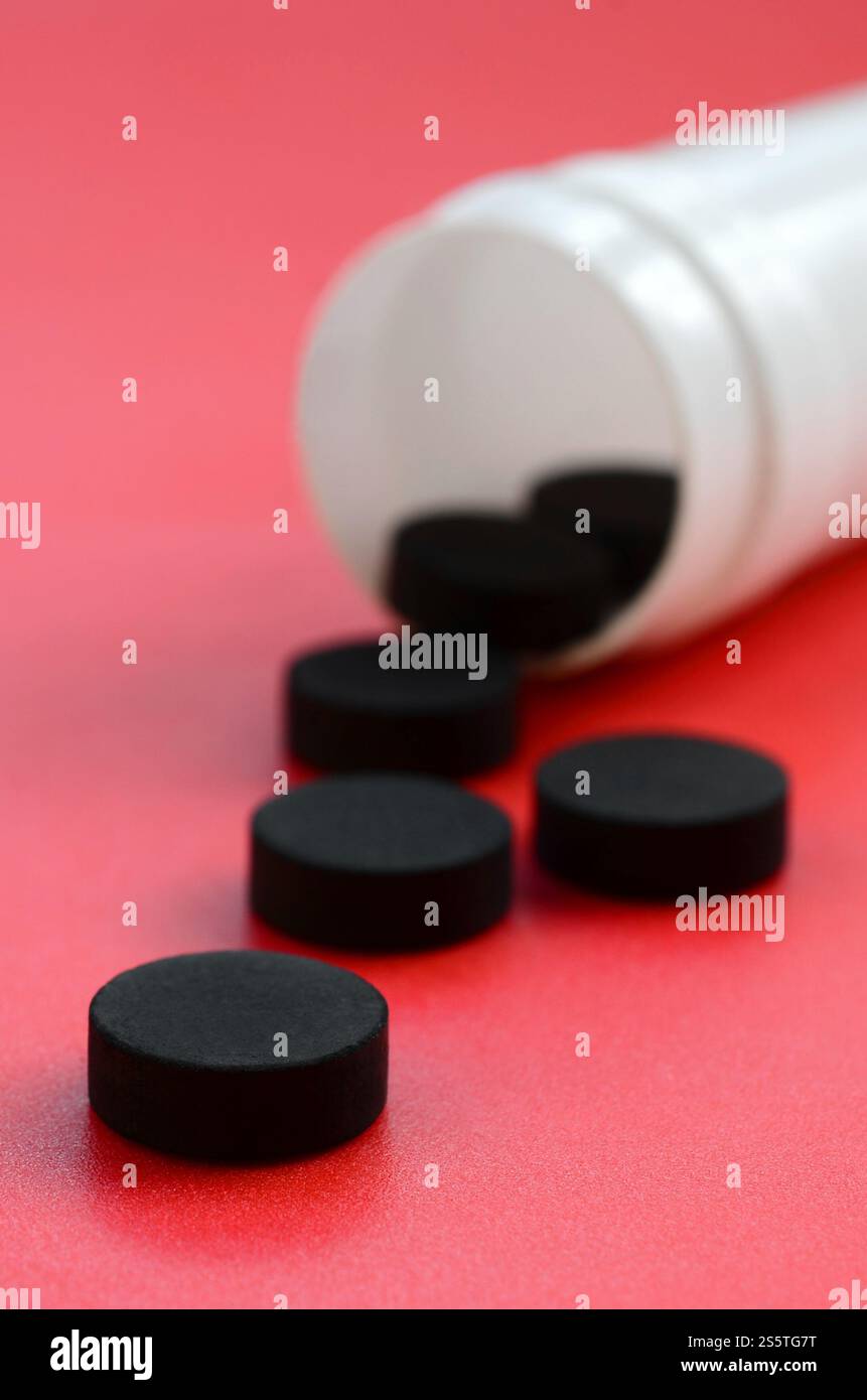 Mehrere schwarze Tabletten fallen aus dem Kunststoffbehälter auf der roten Oberfläche. Hintergrundbild zu medizinischen und pharmazeutischen Themen. Aktivkohle Stockfoto