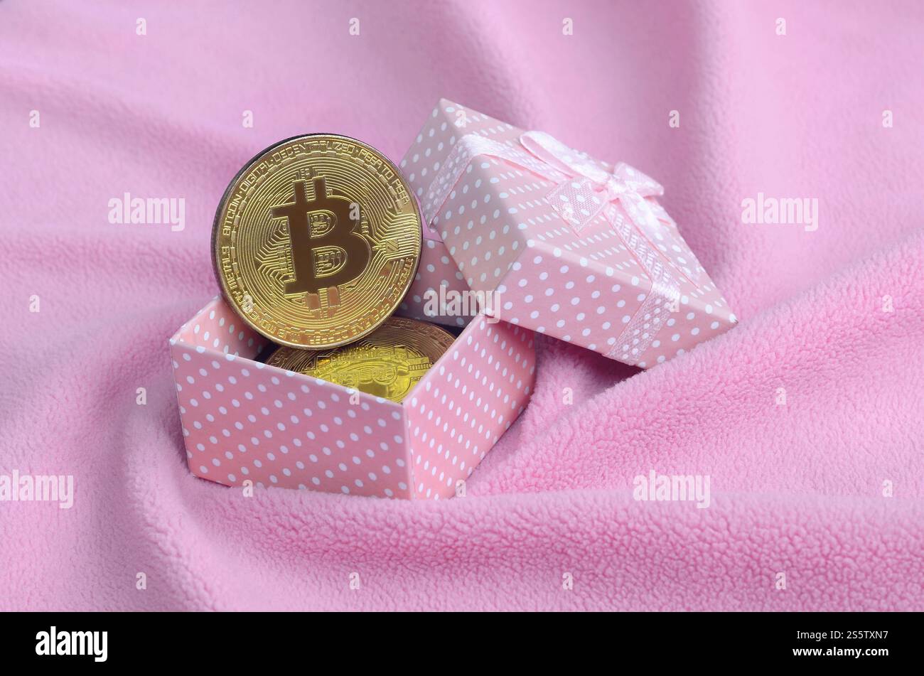 Der goldene Bitcoin liegt in einer kleinen rosafarbenen Geschenkbox mit einer kleinen Schleife auf einer Decke aus weichem und flauschigem hellrosa Fleecestoff mit einer großen Anzahl Stockfoto