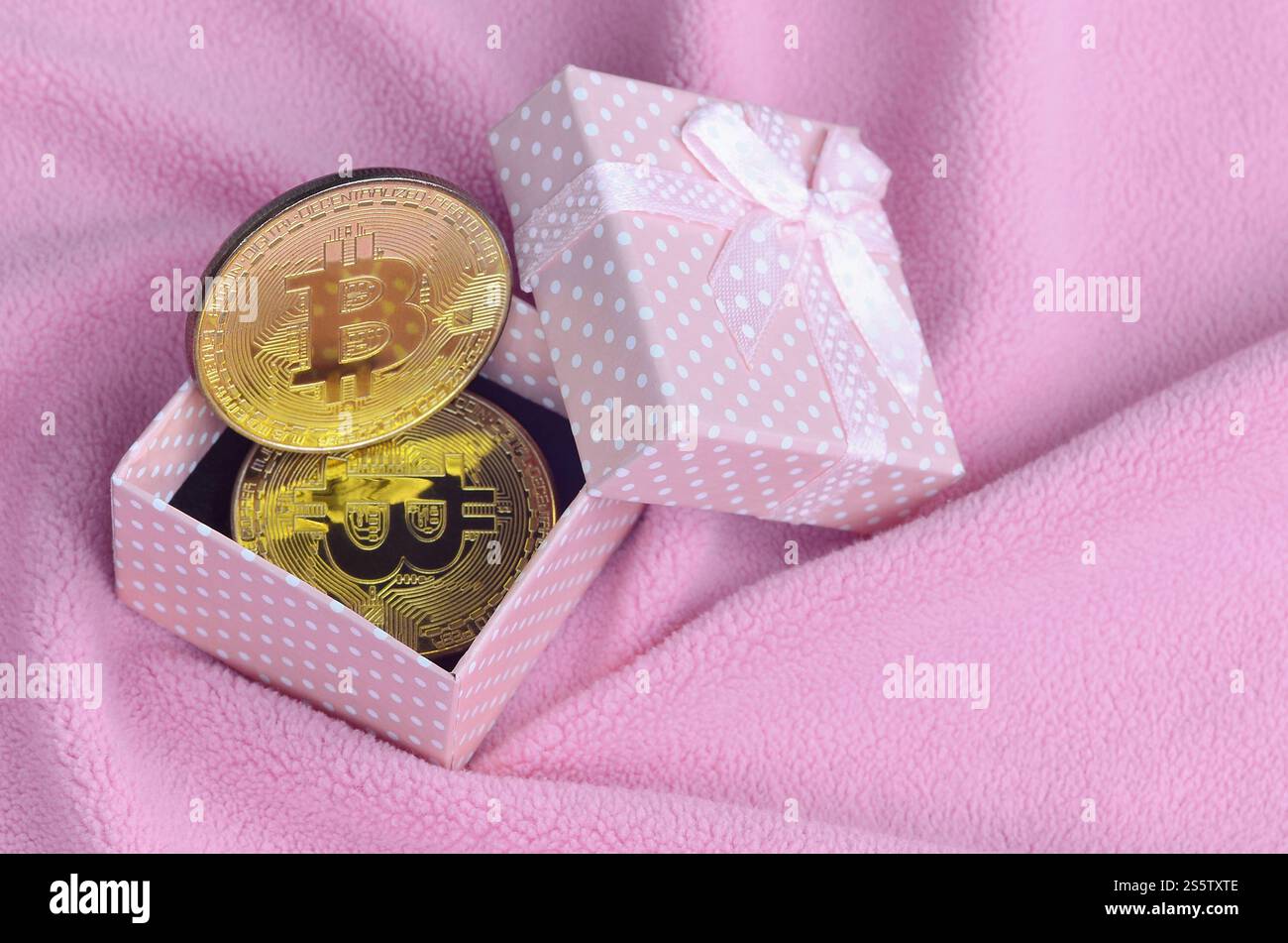 Der goldene Bitcoin liegt in einer kleinen rosafarbenen Geschenkbox mit einer kleinen Schleife auf einer Decke aus weichem und flauschigem hellrosa Fleecestoff mit einer großen Anzahl Stockfoto