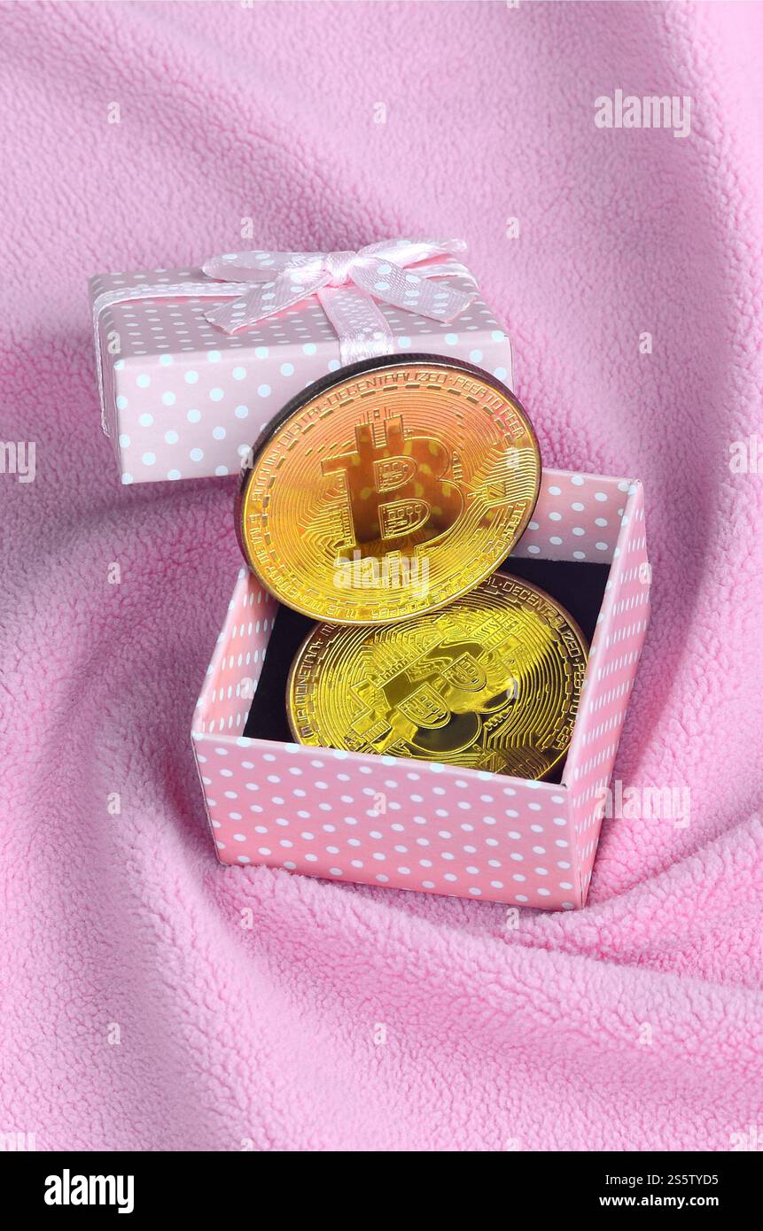 Der goldene Bitcoin liegt in einer kleinen rosafarbenen Geschenkbox mit einer kleinen Schleife auf einer Decke aus weichem und flauschigem hellrosa Fleecestoff mit einer großen Anzahl Stockfoto