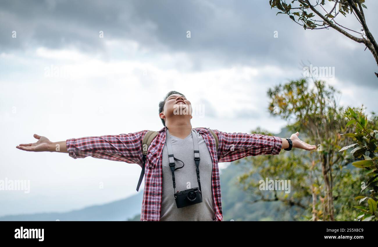 Junger asiatischer Wandermann mit Kamera stehend und aufgerichtet Hände mit glücklich und frisch auf dem Gipfel des felsigen Berges, bewölkt über dem Tal im Hintergrund, Stockfoto