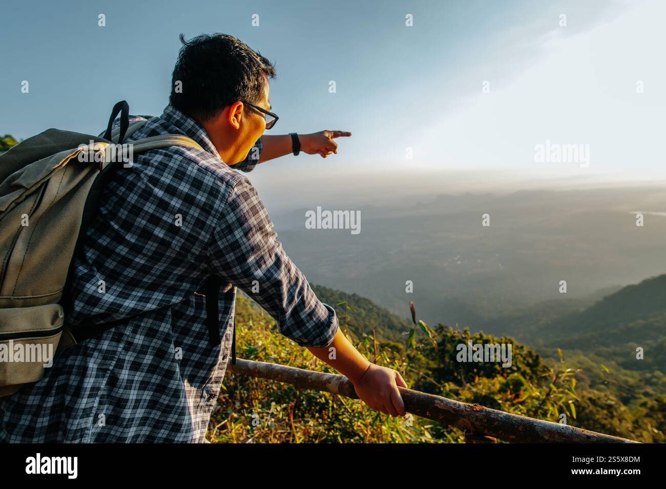 Junger asiatischer Wanderer mit Rucksack stehend und zeigen Sie mit dem Finger nach vorne auf einen wunderschönen Blick auf das Tal am Aussichtspunkt mit Sonnenstrahl bei Sonnenuntergang, Kopierraum Stockfoto