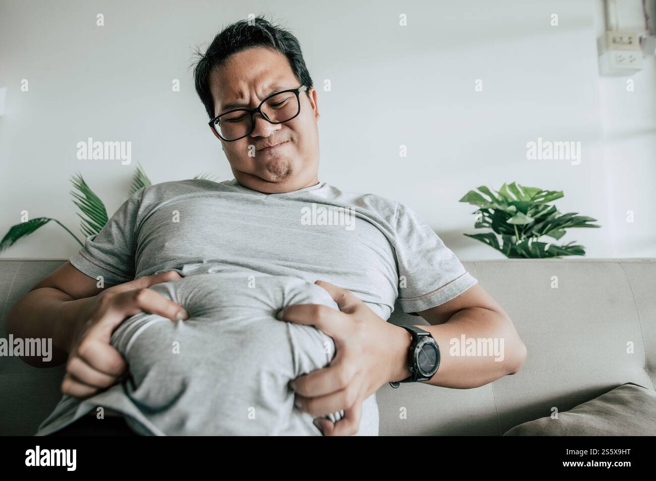 Fetter Mann in Brille sitzt auf dem Sofa und zeigt Adipositas mit Stress, lustiger übergewichtiger Mann usd Hände drücken seinen Bauch fett, Kopierraum Stockfoto