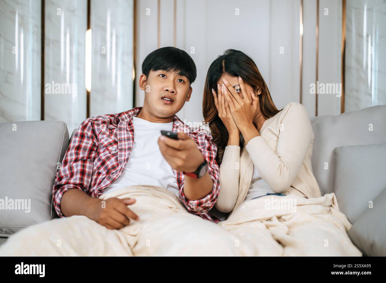 Junge asiatische Paare verbringen nachmittags im Wohnzimmer schaurige Filme im Fernsehen, Junge Mann und Frau sitzen auf dem Sofa und decken sich mit einer Decke ab Stockfoto