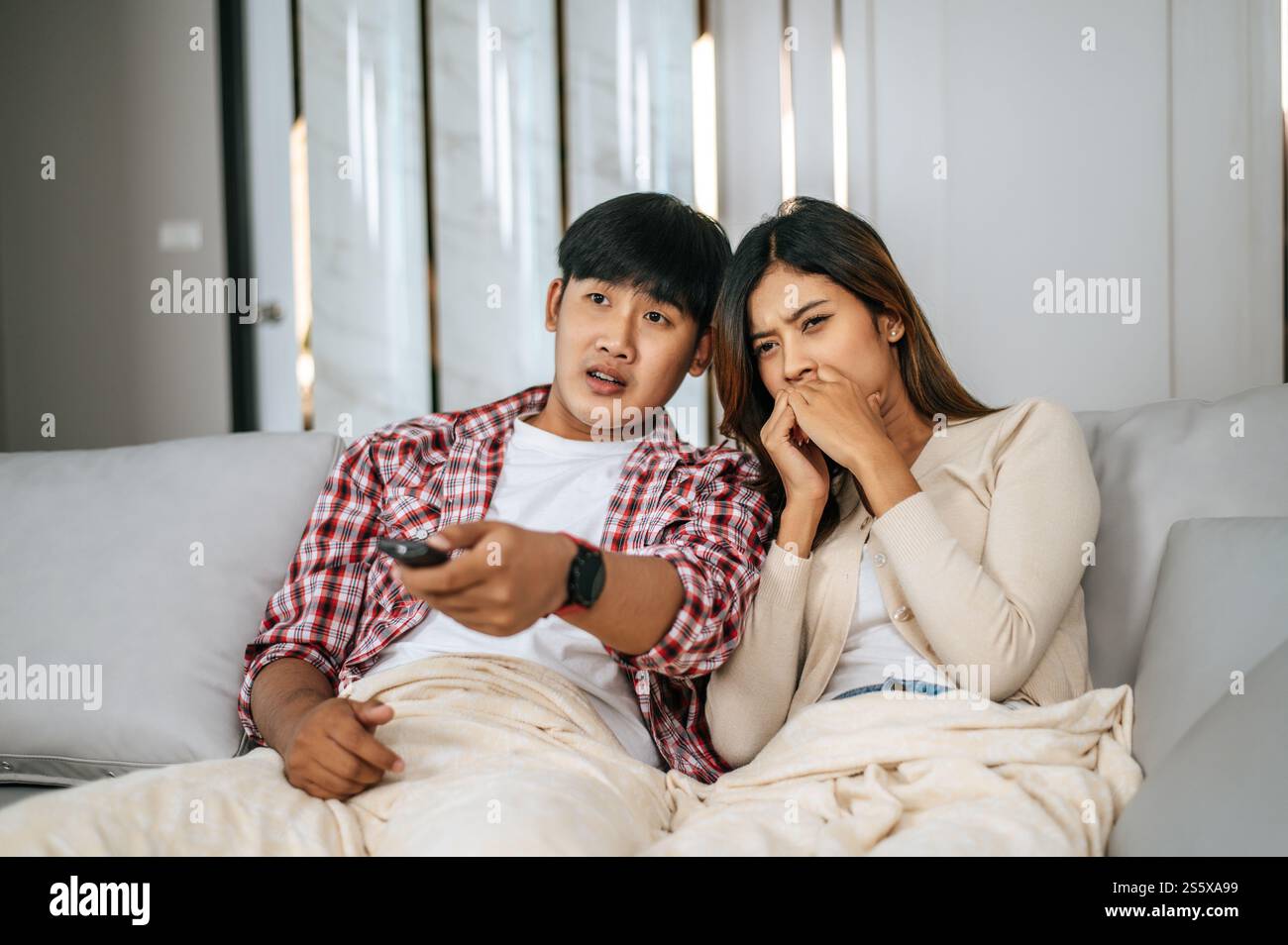Junge asiatische Paare verbringen nachmittags im Wohnzimmer schaurige Filme im Fernsehen, Junge Mann und Frau sitzen auf dem Sofa und decken sich mit einer Decke ab Stockfoto