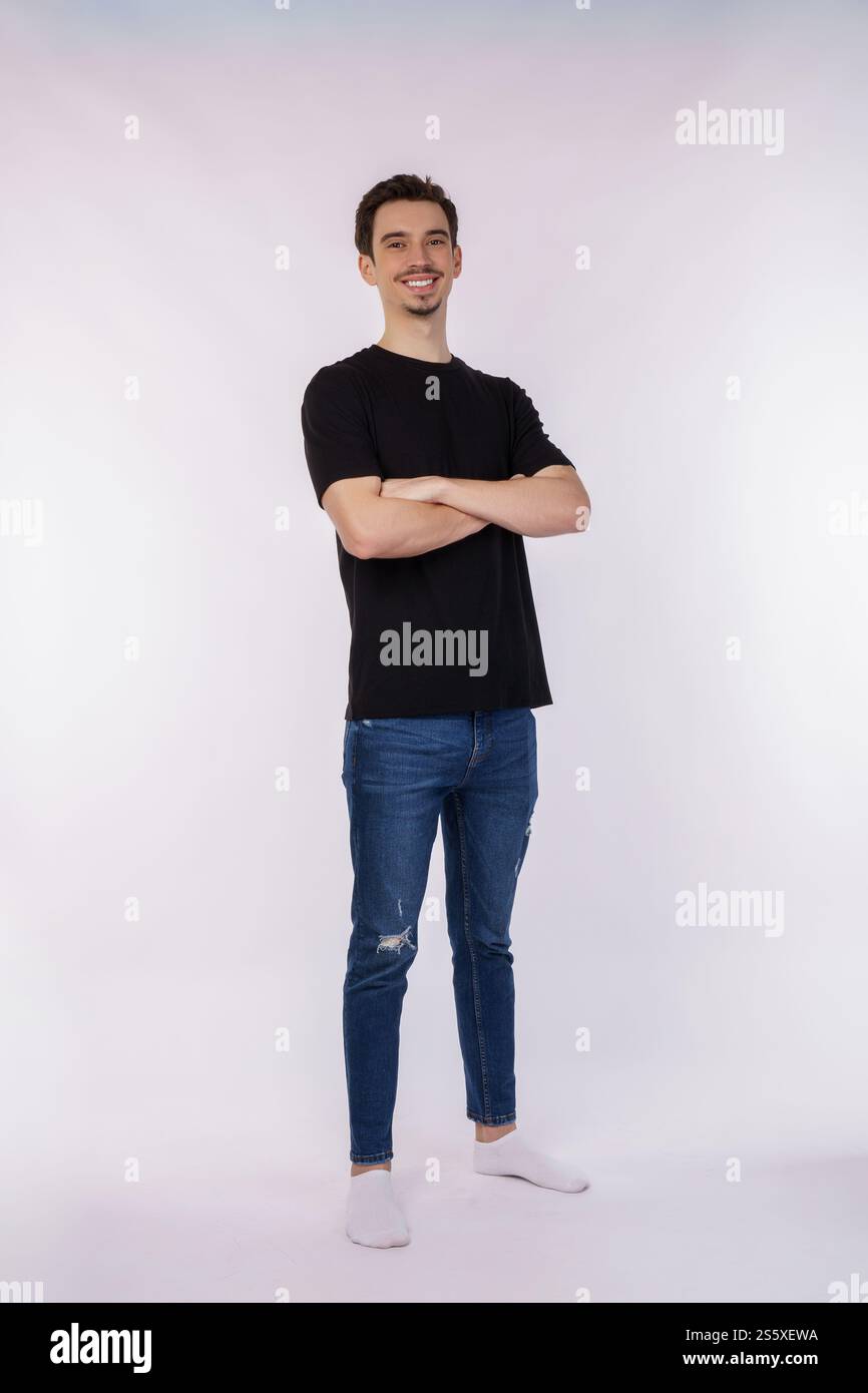 Porträt eines jungen gutaussehenden Mannes, der ein schwarzes T-Shirt trägt, mit überkreuzten Armen mit lokalem weißem Hintergrund im Studio steht. Stockfoto