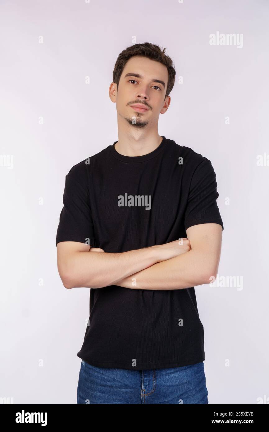 Porträt eines jungen gutaussehenden Mannes, der ein schwarzes T-Shirt trägt, mit überkreuzten Armen mit lokalem weißem Hintergrund im Studio steht. Stockfoto