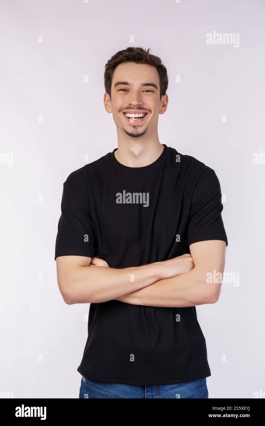 Porträt eines jungen gutaussehenden Mannes, der ein schwarzes T-Shirt trägt, mit überkreuzten Armen mit lokalem weißem Hintergrund im Studio steht. Stockfoto