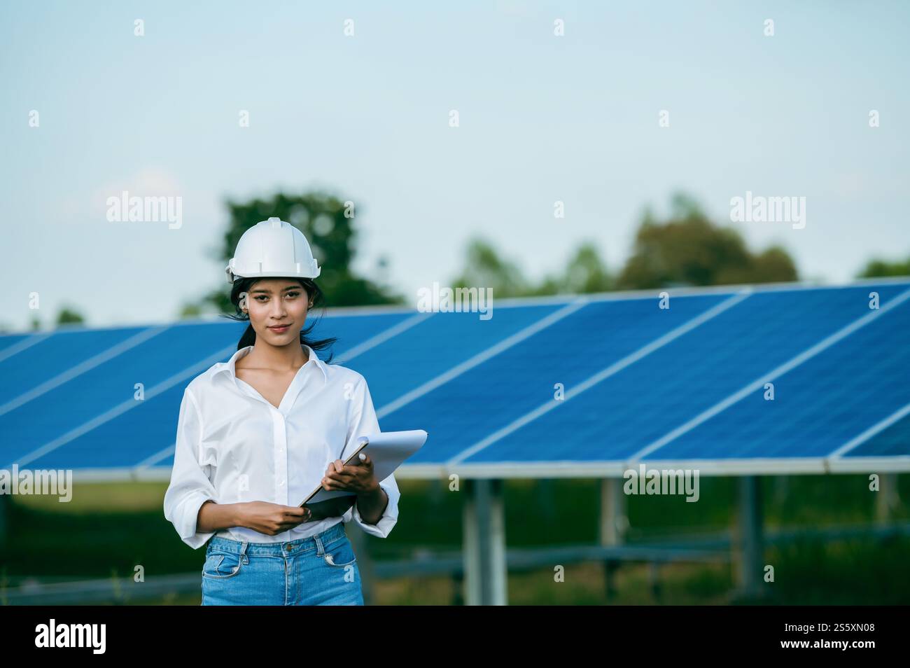 Porträt Asiatische junge Ingenieurin, die einen Schutzhelm trägt, der vor dem Sonnenkollektor steht, um den Betrieb in Papieren zu überprüfen, während sie arbeiten Stockfoto