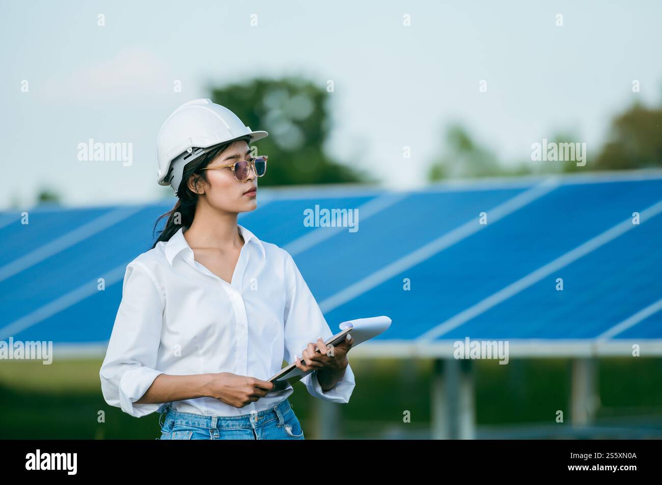 Porträt Asiatische junge Ingenieurin, die Schutzhelm und Sonnenbrille trägt, die vor dem Sonnenkollektor steht, um den Betrieb in Papieren zu überprüfen Stockfoto