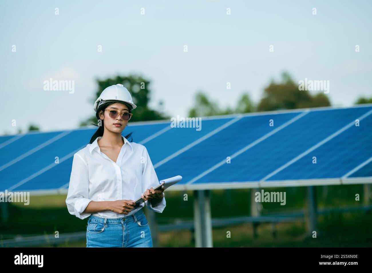 Porträt Asiatische junge Ingenieurin, die Schutzhelm und Sonnenbrille trägt, die vor dem Sonnenkollektor steht, um den Betrieb in Papieren zu überprüfen Stockfoto