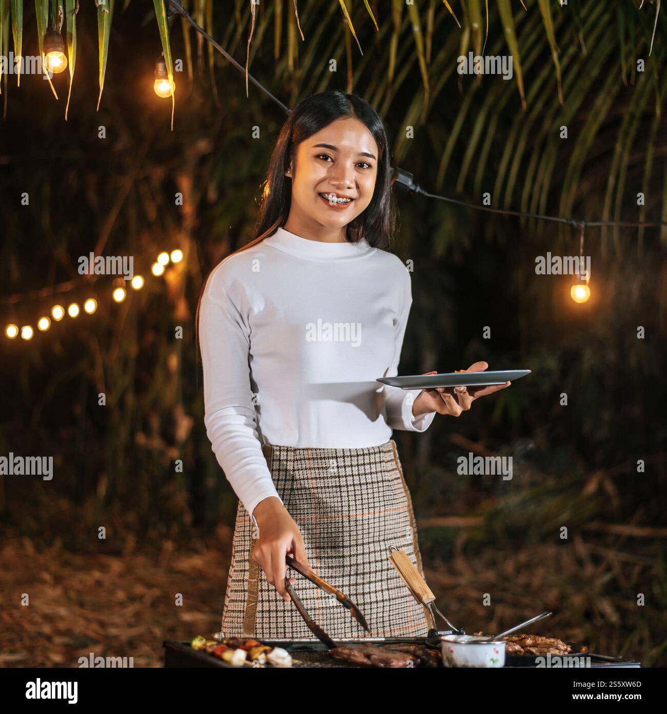 Porträt einer glücklichen asiatischen jungen Frau, die Fleisch auf dem Grill auf der Neujahrsfeier kocht. Bar-B-Q oder BBQ auf traditionellem Herd. Nachtparty, Leute und Stockfoto