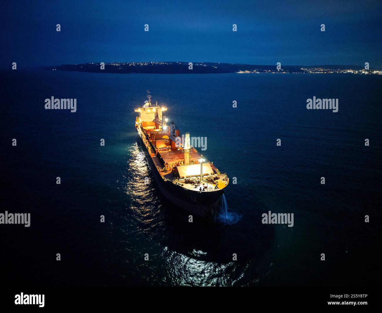 Beleuchtetes Frachtschiff auf See bei Nacht, von oben gesehen mit Küste und leuchtenden Lichtern der Stadt in der Ferne Stockfoto