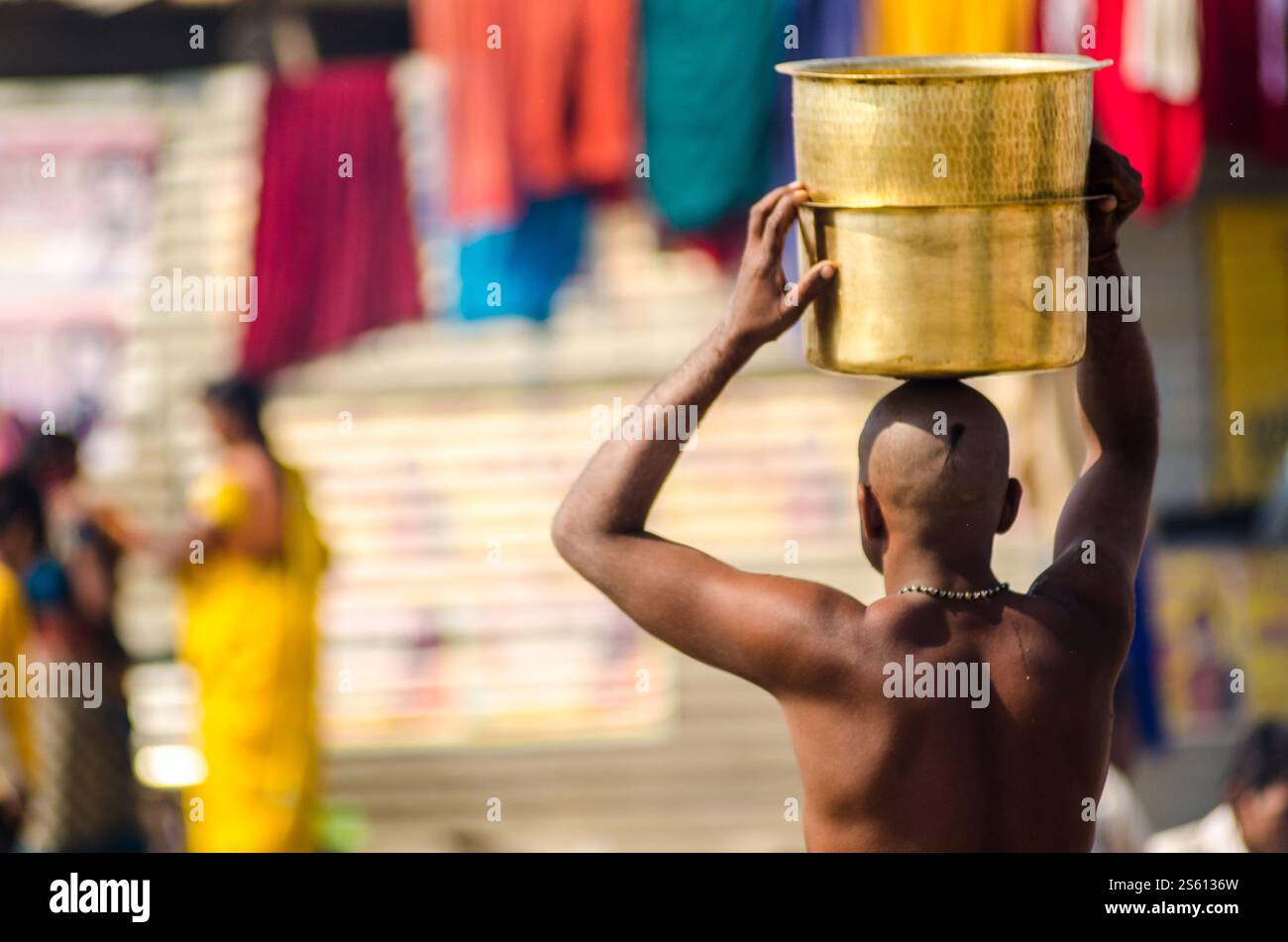 Die Person trägt einen goldenen Topf auf dem Kopf, während sie durch einen pulsierenden Markt in einem belebten Stadtgebiet spaziert Stockfoto