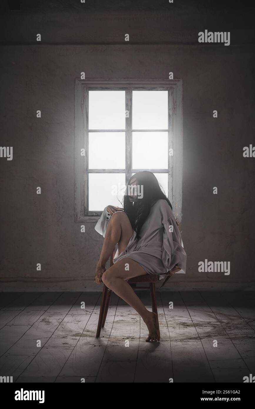 Eine gruselige Frau mit schwarzem Haar, weißer Haut und dunklen Augen sitzt auf dem Stuhl und gewinnt ein weißes Kleid in einer furchterregenden Haltung, das das Gefühl des Grauens und weckt Stockfoto