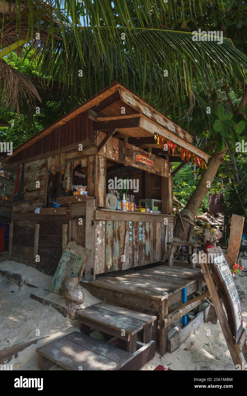 Eine verwitterte hölzerne Strandhütte mit der Bezeichnung Love Bar befindet sich unter üppigem Laub an einem Sandstrand, umgeben von Palmenwedeln und schiefen Bäumen in Thailand. Stockfoto