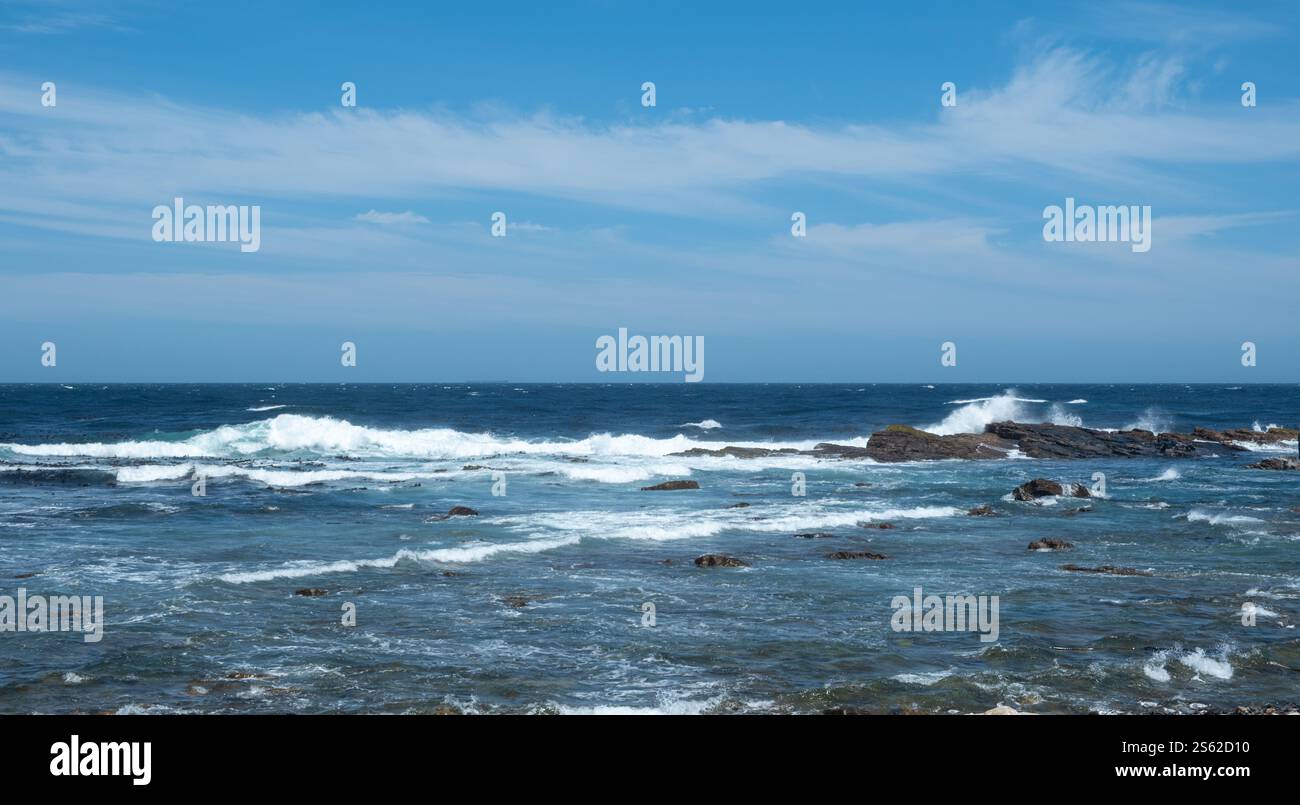 Küstenszene, sanfte Meereswellen, die gegen felsiges Ufer brechen, unter einem leuchtend blauen Himmel mit verstreuten Wolken. Stockfoto