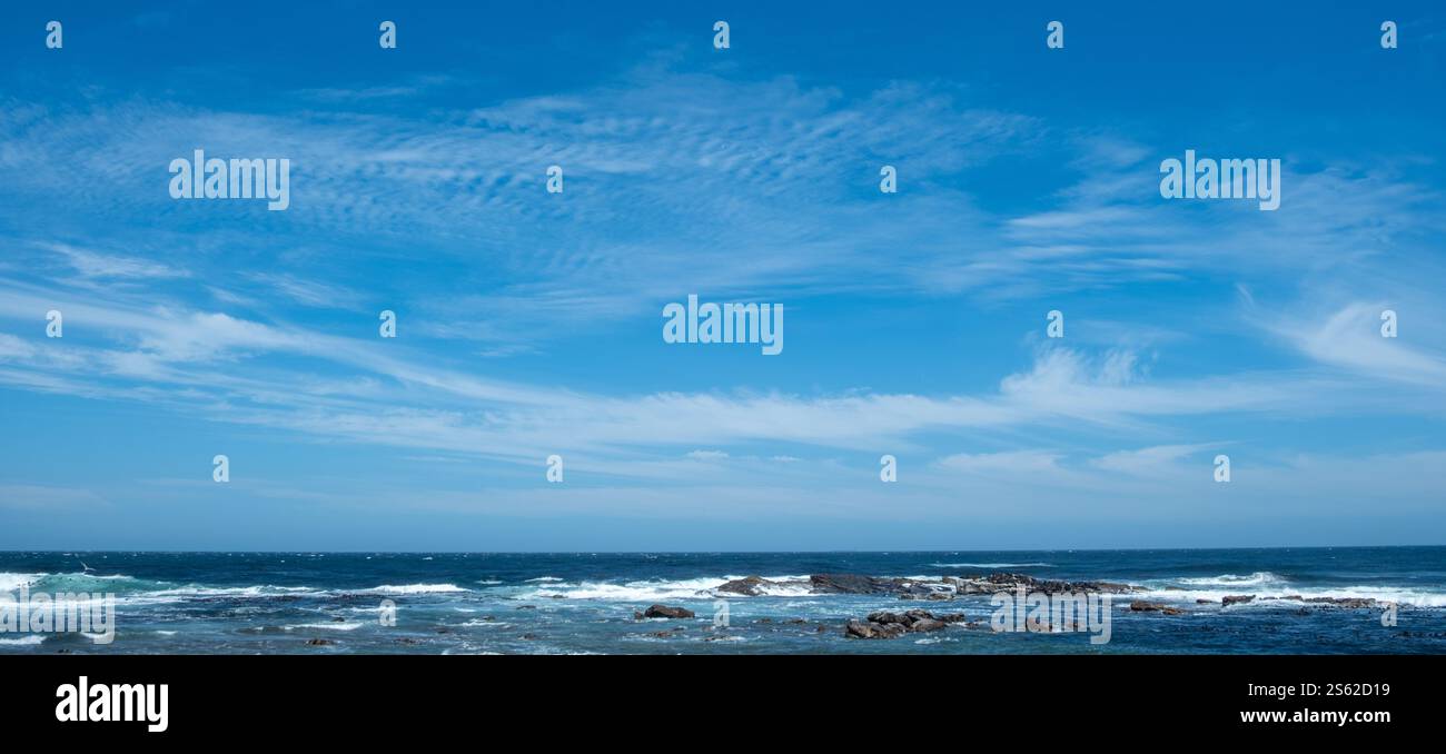 Küstenszene, sanfte Meereswellen, die gegen felsiges Ufer brechen, unter einem leuchtend blauen Himmel mit verstreuten Wolken. Stockfoto