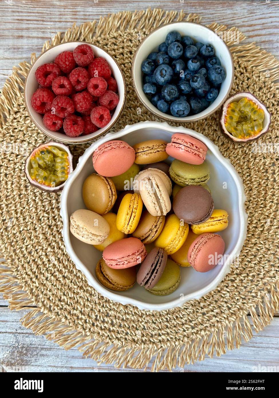 Macarons in verschiedenen Farben und Geschmacksrichtungen und Himbeeren, Heidelbeeren und Passionsfruchthälften auf der Korbserviette - Smartphone-aufgenommenes Stockfoto