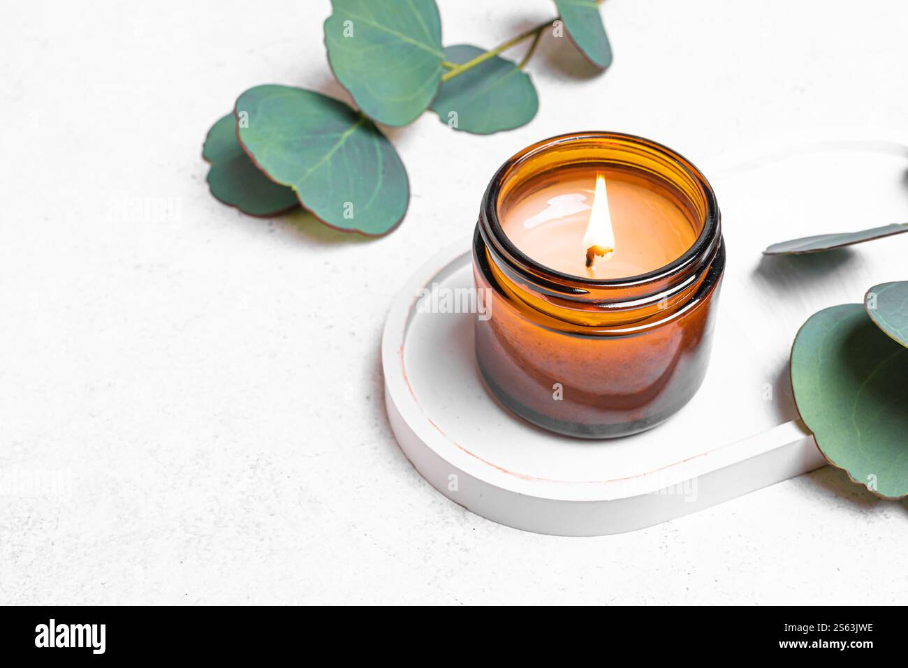 Brennende Soja-Wachskerze in bernsteinfarbenem Glas und grüne eucaliptus-Blätter auf weißem Betontablett. Natürliches Wohndekor, Spa-Vorlage und Aroma-Massage-Conce Stockfoto