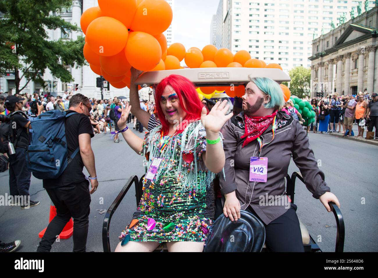 Teilnehmer in lebhaften Kostümen und Accessoires fahren während eines lebhaften Pride Festivals auf einem mit orangen Ballons geschmückten Wagen Stockfoto