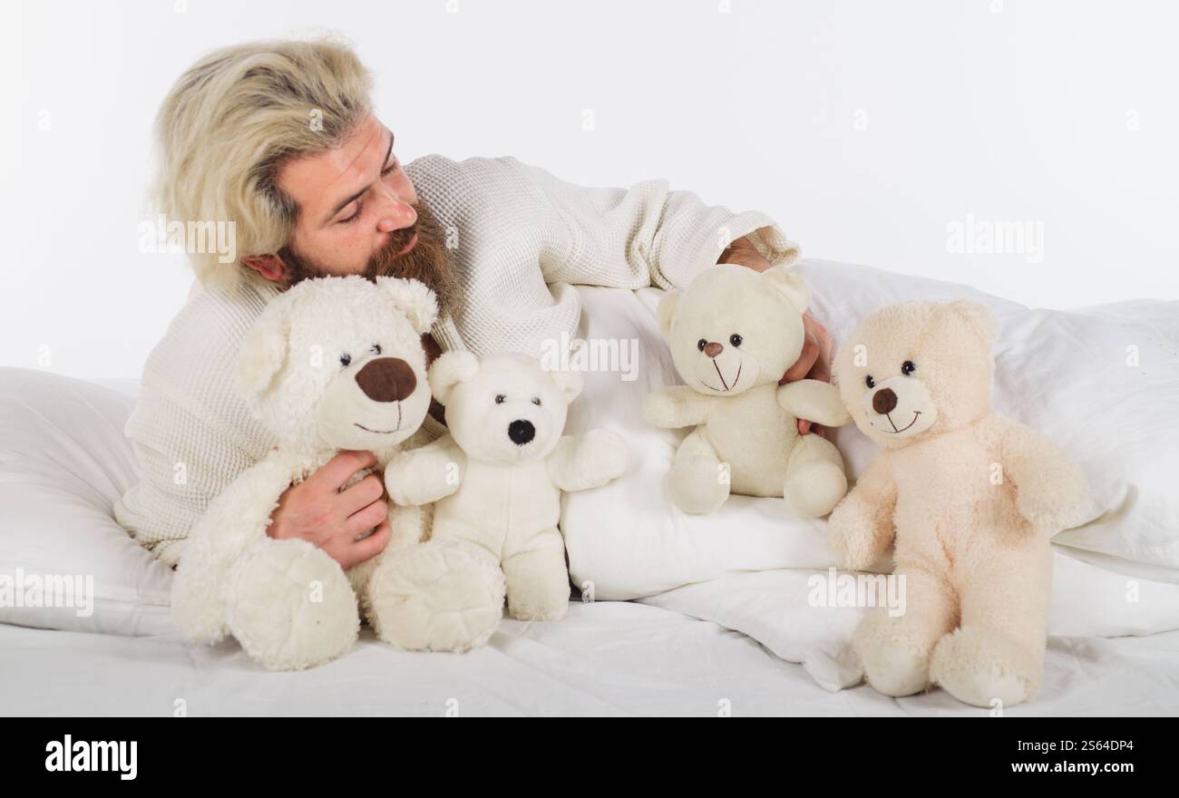 Lächelnder bärtiger Mann, der mit vielen Teddybären im Bett liegt. Guten Morgen. Hübscher Kerl in einem bequemen Bett auf weißer Bettwäsche mit Plüschspielzeug. Schläfriger Mann in Stockfoto
