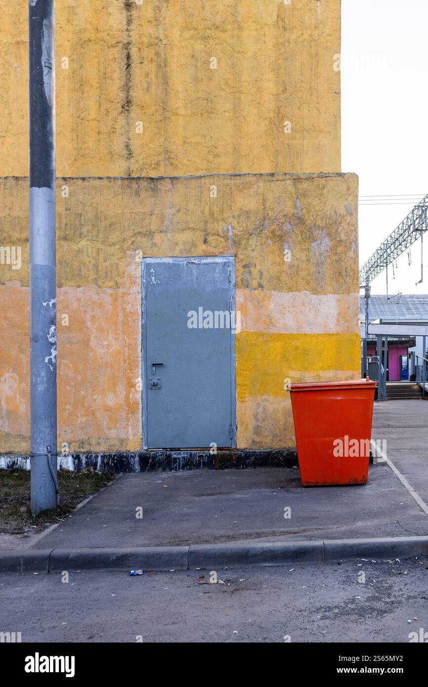 Schäbig lackierte gelbe Wand des Industriegebäudes, roter Straßenmüll neben dem Bahnsteig am bewölkten Herbsttag Stockfoto