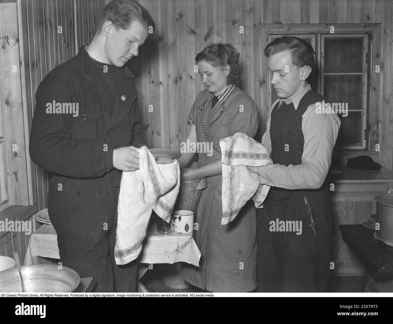 Während des Zweiten Weltkriegs. Der Krieg erschwerte den Import von Kohle für Heizhäuser und Wohnungen. Statt Kohle musste Holz verwendet werden. In Großstädten wie Stockholm konnte man große Holzhaufen entlang der Straßen sehen. Viele Männer erhielten befristete Jobs, um am Holzhacken zu arbeiten. Hier sind zwei junge Männer, die in den Wäldern außerhalb der Västmanlandstadt Skinnskatteberg arbeiten. Sie machen eine dringend benötigte Pause in ihrer einfachen Hütte, wo es keinen Strom gibt. Eine junge Frau wird als Köchin angestellt, wird aber in der Küche von zwei Männern unterstützt, die eifrig das Geschirr trocknen. Wahrscheinlich Stockfoto