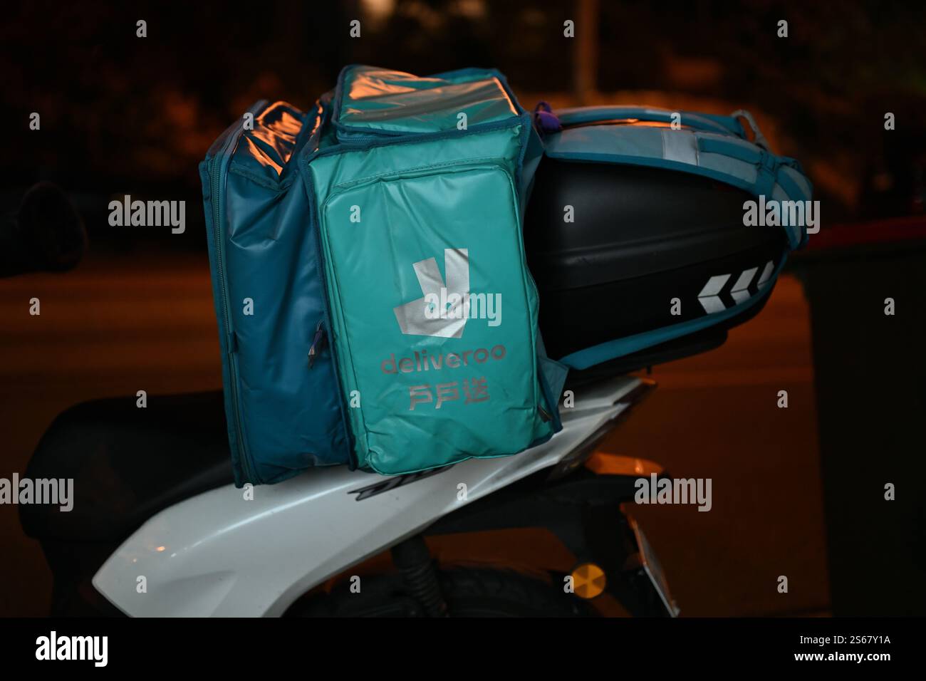 Seitenansicht einer Deliveroo Aufbewahrungstasche mit Firmenlogo und chinesischen Schriftzeichen auf der Rückseite eines geparkten Motorrollers bei Nacht Stockfoto