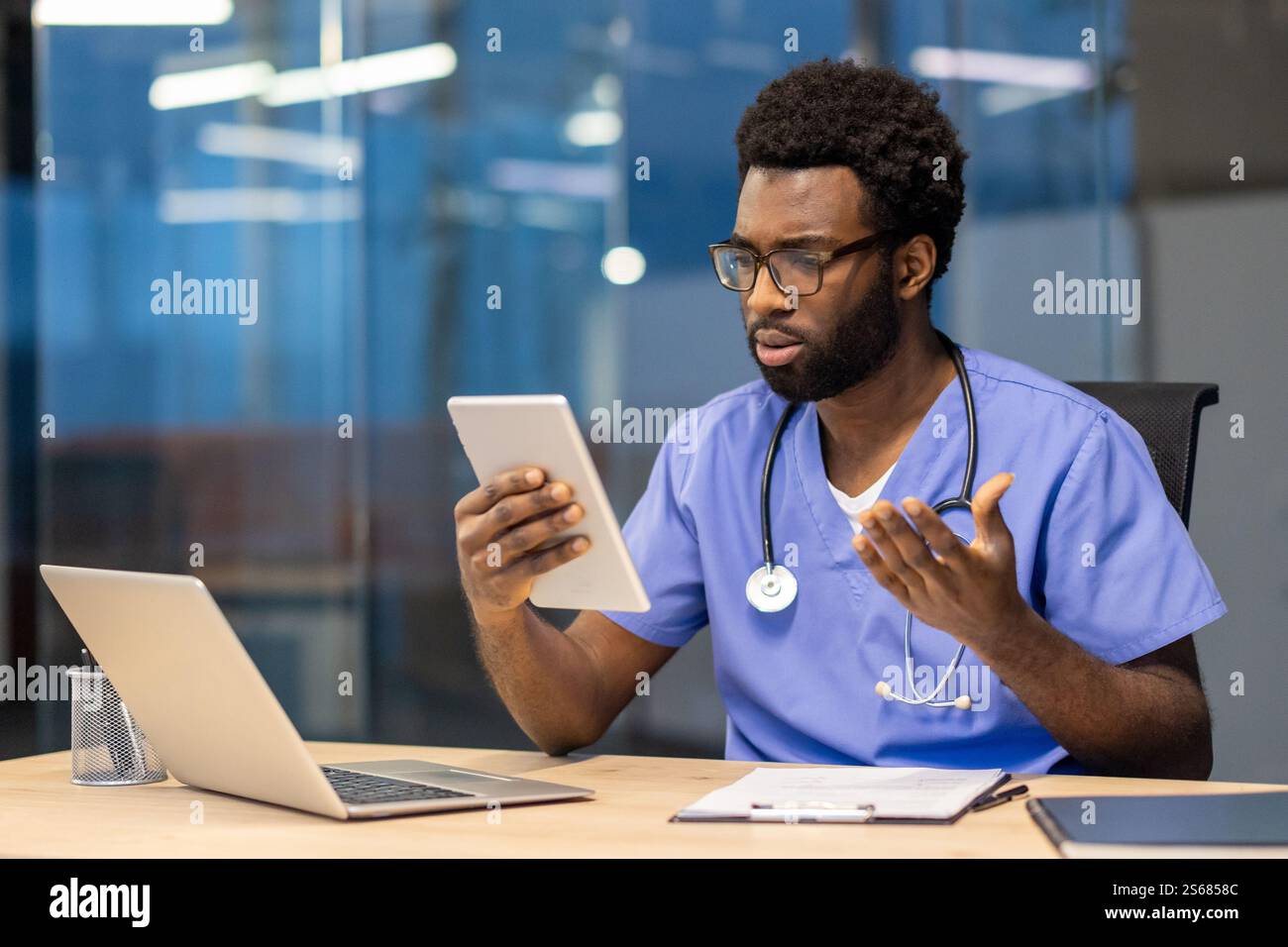 Junger erfahrener Arzt mit Tablet-Computer, der am Schreibtisch sitzt, mit Kollegen spricht und Patienten aus der Ferne konsultiert. Mann in blauem medizinischem Mantel und Stethoskop, der in der Klinik sitzt. Stockfoto
