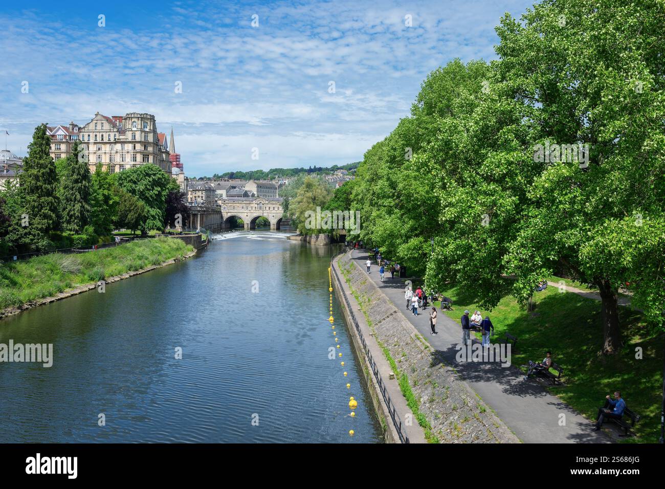 Menschen, die entlang des Flusses Avon in Bath UK spazieren, mit der historischen Pulteney Bridge und dem Empire Hotel, die eine atemberaubende architektonische Kulisse bieten. Stockfoto