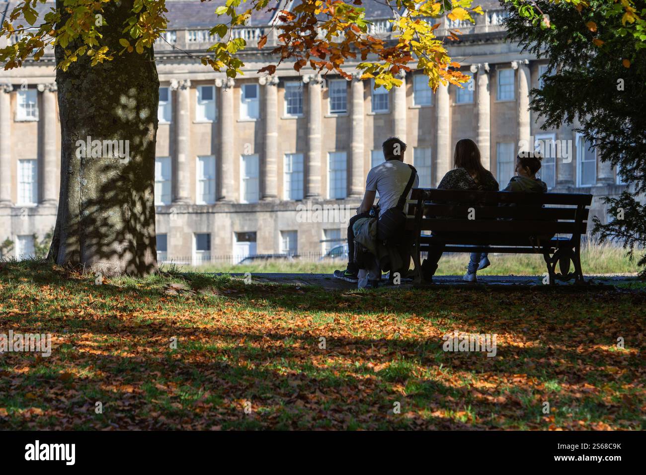 Die Menschen sitzen auf einer Bank vor dem historischen Circus in Bath, Großbritannien, umgeben von georgianischer Architektur und einer friedlichen urbanen Umgebung. Stockfoto