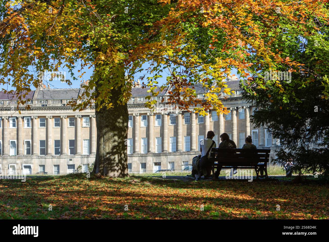 Die Menschen sitzen auf einer Bank vor dem historischen Circus in Bath, Großbritannien, umgeben von georgianischer Architektur und einer friedlichen urbanen Umgebung. Stockfoto