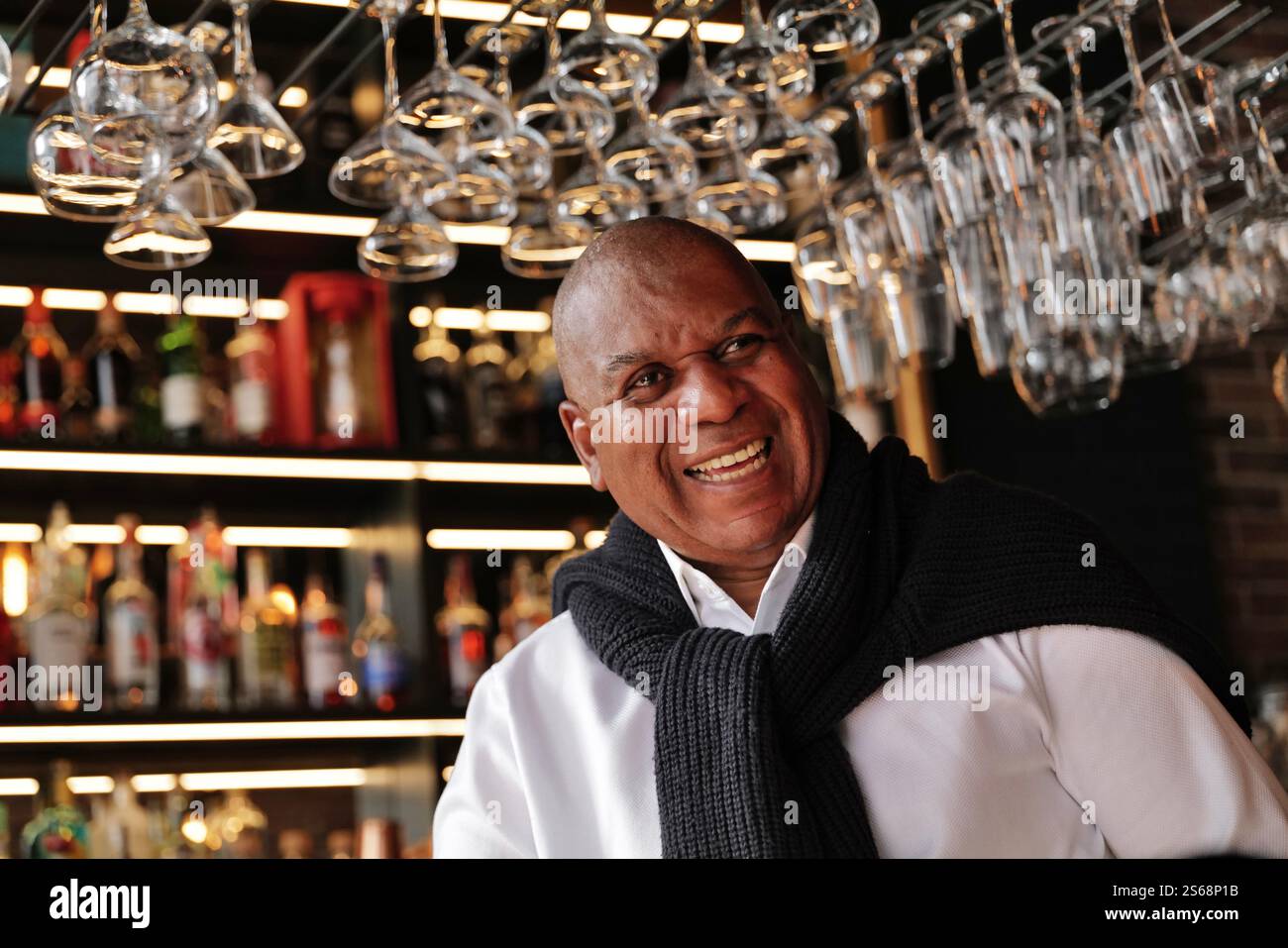 Ein glücklicher schwarzer Seniorenmann steht an der Bar, umgeben von Flaschen und Gläsern in einer trendigen Bar. Stockfoto Ein glücklicher schwarzer Seniorenmann steht an der Bar, umgeben von Flaschen und Gläsern in einer trendigen Bar. Stockfoto
