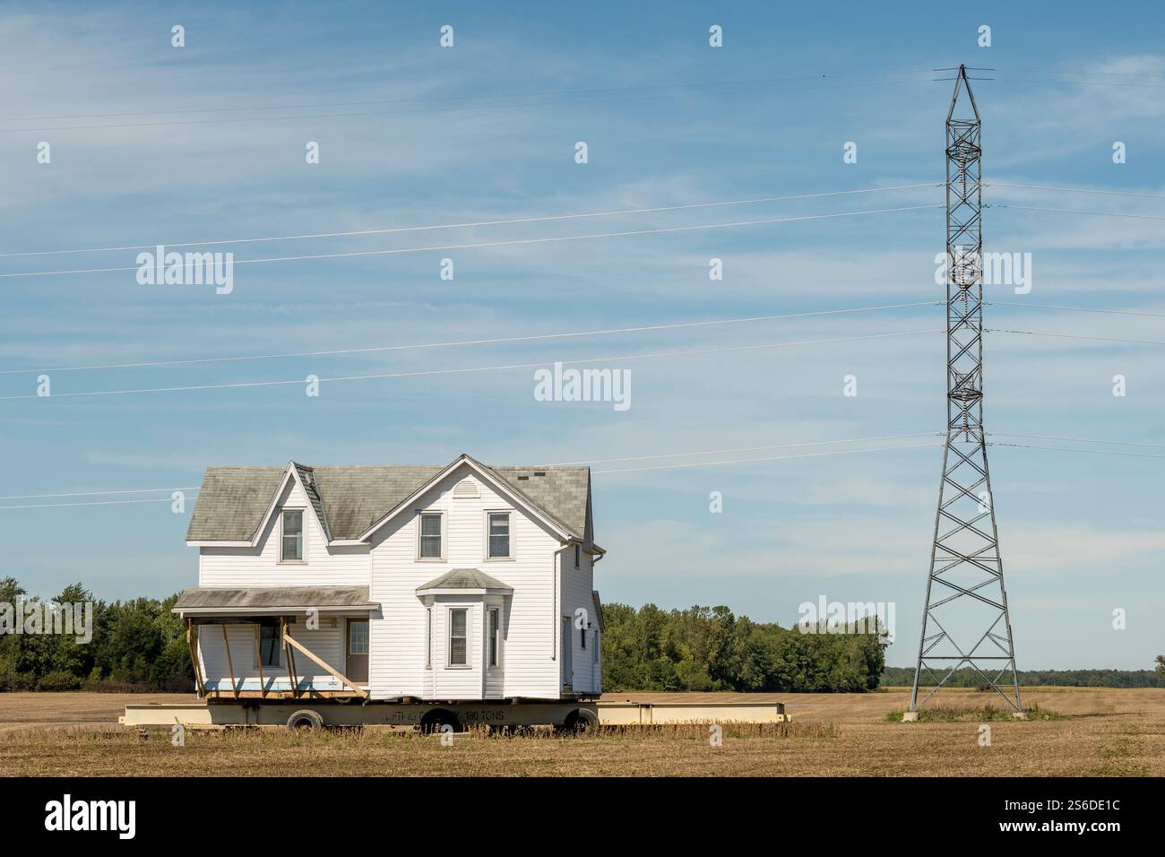 Weißes Bauernhaus wird über ein Bauernfeld transportiert, auf einem Anhänger unter einem hellblauen Himmel balanciert, mit einer hoch aufragenden Übertragungsleitung in der Nähe. Stockfoto