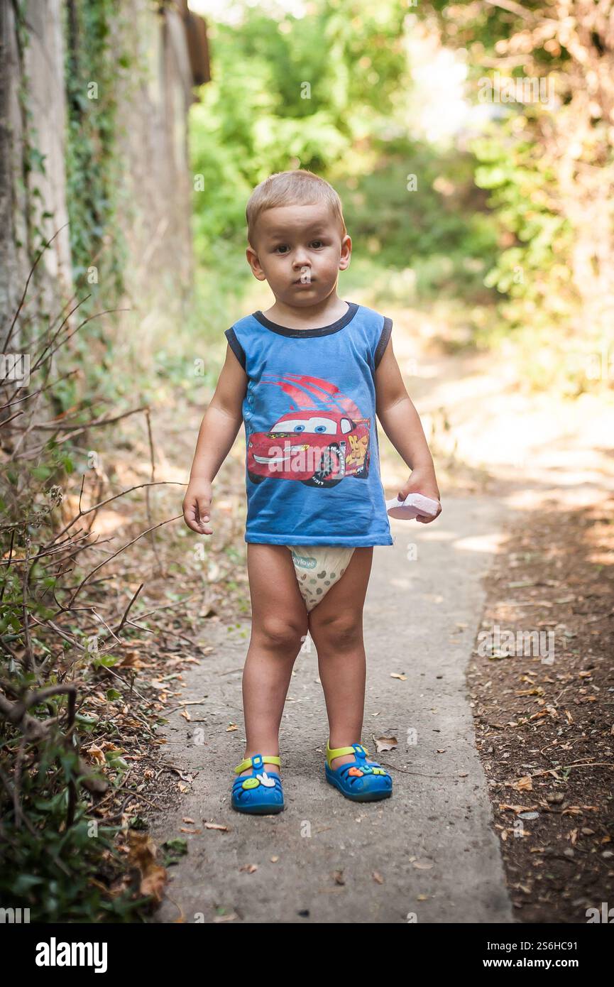 Ein kleines Kind spaziert auf einem schmalen Weg, umgeben von Grün, gekleidet mit einem Tanktop und einer Windel. Helles Sonnenlicht filtert durch Bäume und erzeugt einen Stockfoto
