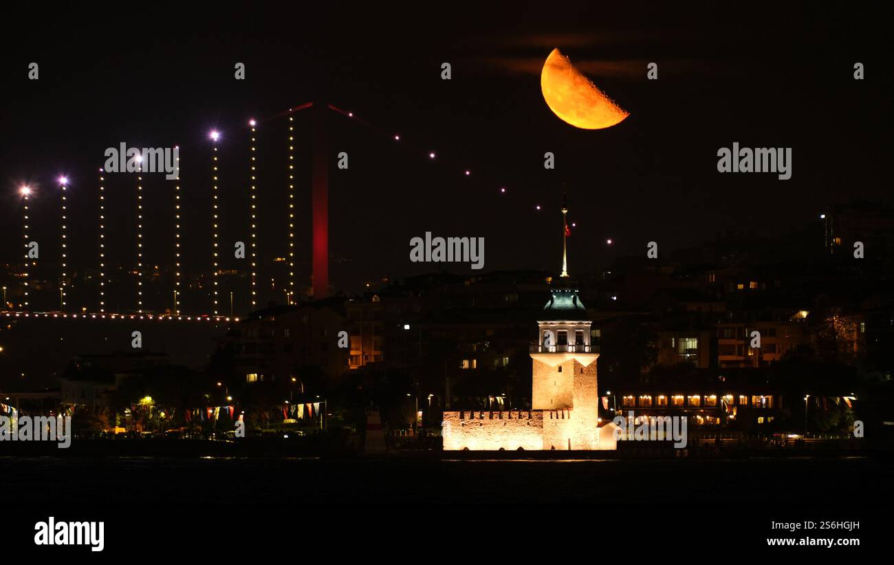 Ein atemberaubender Blick bei Nacht auf Istanbul mit dem beleuchteten Maidenturm, der Bosporus-Brücke und einem leuchtenden orangefarbenen Halbmond. Stockfoto