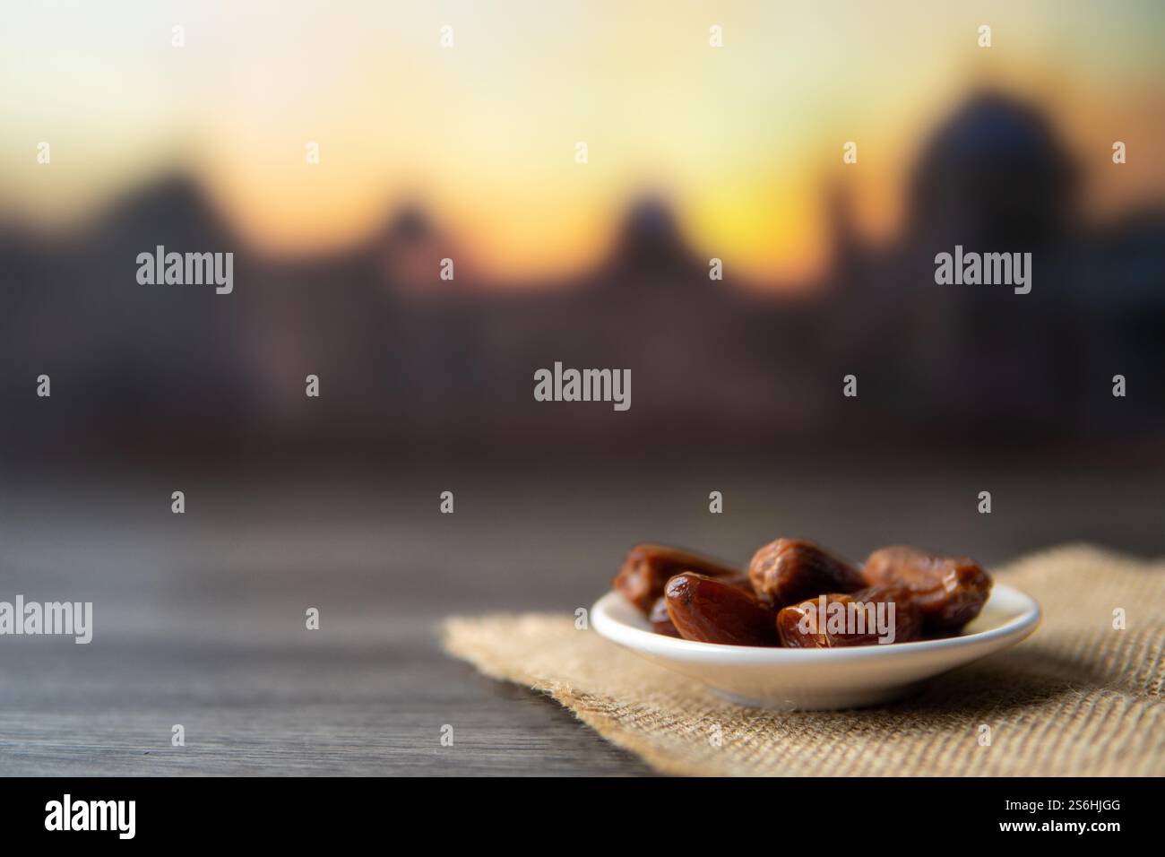 Eine Schüssel Datteln auf einem Sacklappen vor dem Hintergrund eines Sonnenuntergangs und einer Moschee, die den Geist des Ramadan und der islamischen Kultur weckt. Kopierbereich für Text. Stockfoto
