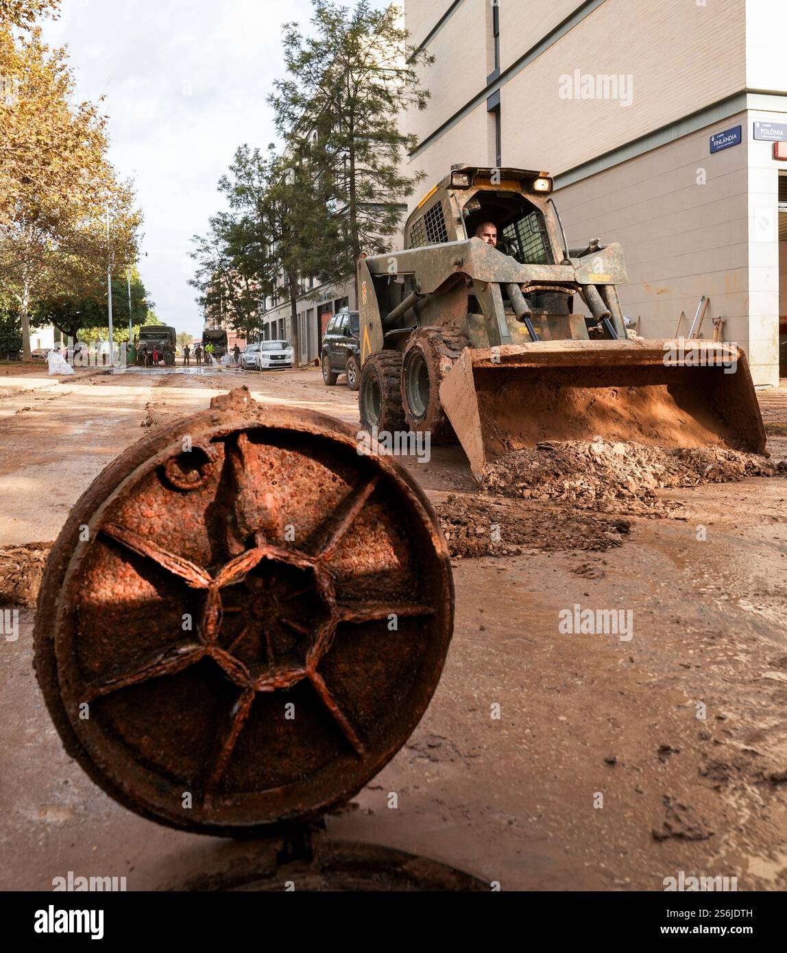 Minibagger-Bobcat und Fahrer, der sich an Schlamm mit Mannlochabdeckung im Vordergrund erinnert. Paiporta, Spanien Stockfoto