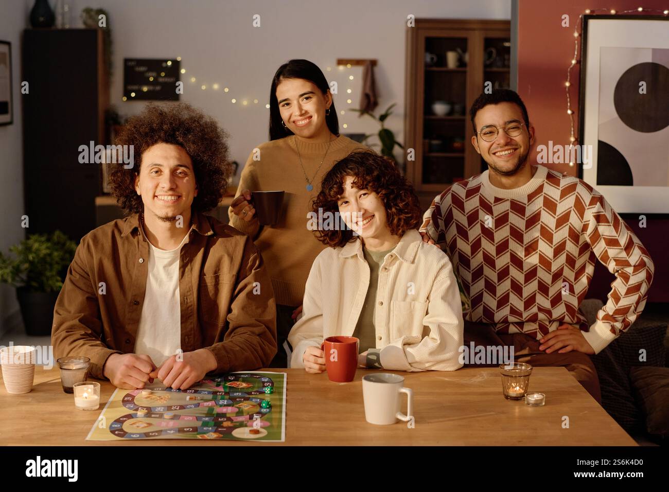 Hochwinkelporträt einer birassischen Gesellschaft von Freunden, die mit Brettspiel und Aromakerzen am Tisch sitzen und die Kamera betrachten, während sie für Gruppenfotos posieren Stockfoto