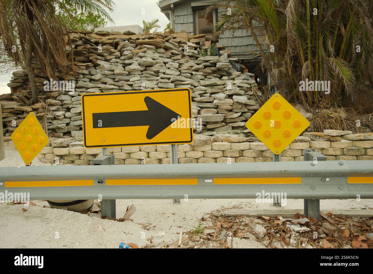 Gelber und schwarzer Pfeil zum rechten Straßenschild mit einem Geländer davor. Großer Stapel flacher Steine und Haus hinten. Ende der Straßenschilder und gr Stockfoto