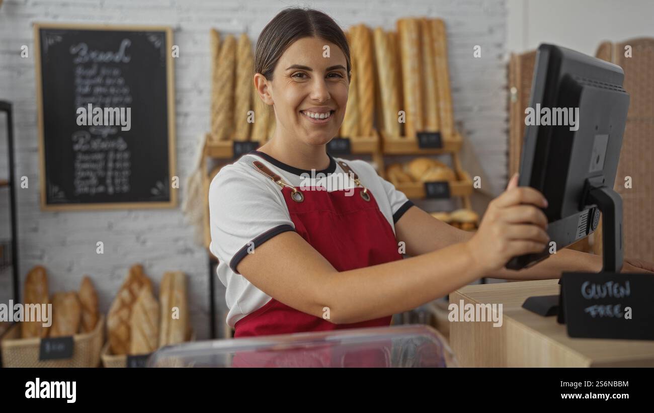 Eine junge Frau mit brünetten Haaren arbeitet in einer Bäckerei mit roter Schürze, umgeben von verschiedenen Brotsorten in einem Innengeschäft. Stockfoto
