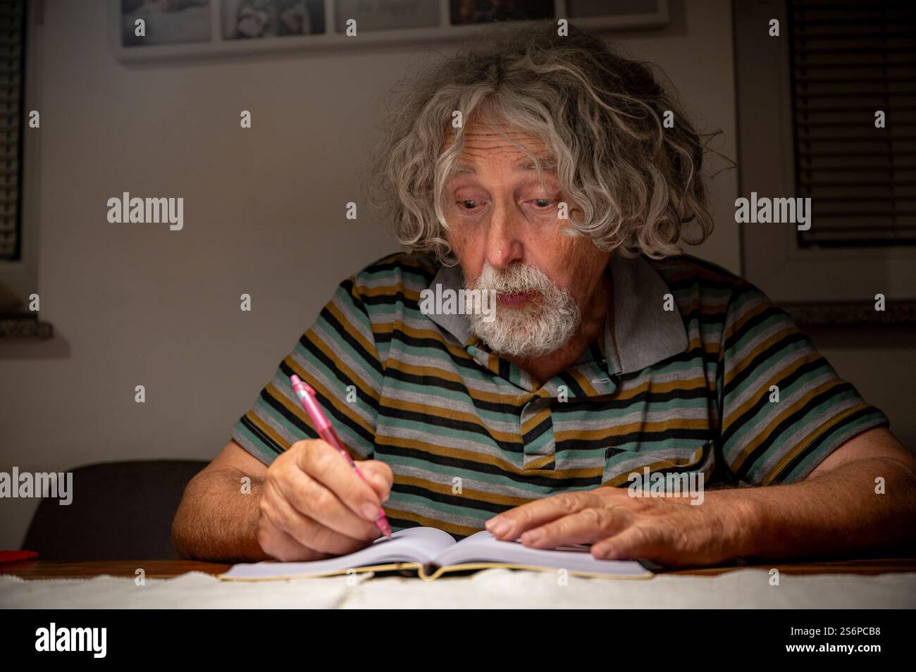 Ein älterer Mann mit lockigen grauen Haaren und einem gestreiften Hemd konzentriert sich tief, während er in einem Notizbuch unter warmer Innenbeleuchtung schreibt. Stockfoto