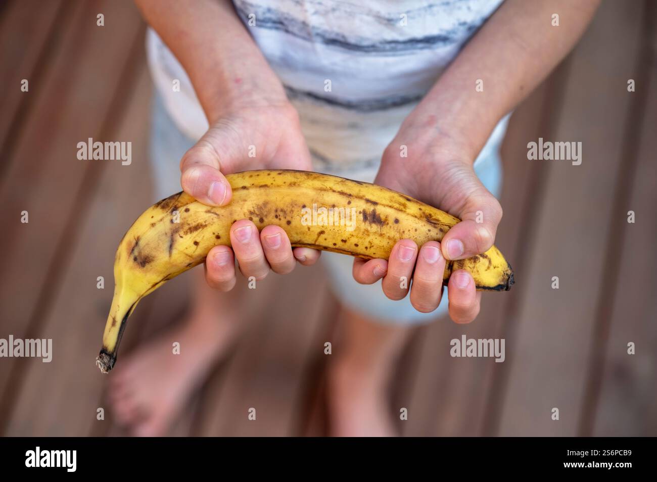 Nahaufnahme der Hände eines Kindes, das sanft eine reife, gefleckte Banane hält und ihre natürlichen Mängel vor einem hölzernen Hintergrund zeigt. Stockfoto