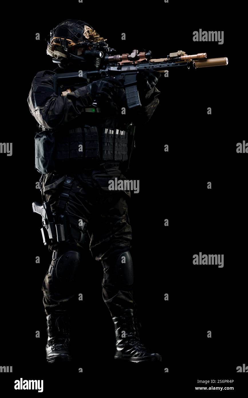 Ein taktischer Soldat in schwarzer Ausrüstung hält ein Gewehr und fokussiert sich auf ein Ziel in einer dunklen Umgebung. Seine Ausrüstung, einschließlich Helm und Kniepolster, hebt r Stockfoto